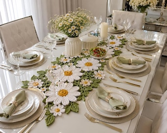 May include: A long, rectangular crochet table runner featuring a repeating pattern of white daisy flowers with yellow centers and green crocheted leaves. The runner is centered on a white dining table set with plates, napkins, and glassware.