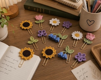 May include: A collection of gold-toned metal paper clips, each topped with a small, crocheted floral or leaf embellishment. The designs include sunflowers, pink tulips, white daisies, purple flowers, blue bell-shaped flowers, and green leaves.