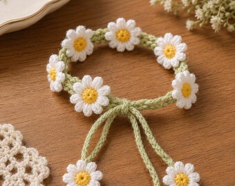 May include: A handmade crochet daisy chain headband. The headband features white daisies with yellow centers, attached to a green crocheted band. The ends of the band have long green ties. The headband is a charming accessory.