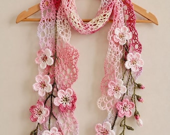 Puede incluir: Bufanda de ganchillo hecha a mano en tonos rosa y blanco, adornada con delicados adornos florales. La bufanda se muestra en una percha de madera, mostrando su intrincado diseño calado y sus acentos florales. Las dimensiones de la bufanda no son visibles.