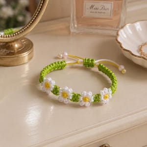 May include: A green and white daisy chain bracelet. The bracelet features a woven green band with white daisy flowers and yellow centers. The bracelet is adjustable with white and yellow cords and small pearl beads.