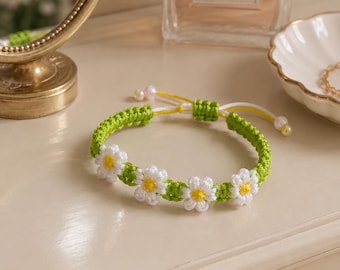 May include: A green and white daisy chain bracelet. The bracelet features a woven green band with white daisy flowers and yellow centers. The bracelet is adjustable with white and yellow cords and small pearl beads.