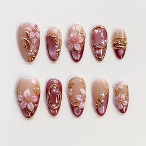 May include: A set of ten almond-shaped artificial nails. Each nail features a gradient design with a nude base and a deep red tip. Embellishments include gold accents, pearls, and pink floral designs.