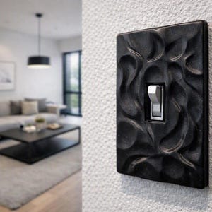 May include: A rectangular black light switch plate featuring a textured, wavy, organic relief pattern. The plate is mounted on a white wall and contains a single, centered, light gray toggle switch with the word ON printed on the switch lever.