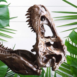 May include: A glossy brown 3D printed Tyrannosaurus rex skull model featuring an articulated jaw that is shown in an open position, revealing detailed teeth and the hollow structure of the cranial cavity and the lower jaw bone.