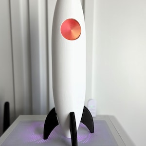 May include: A white rocket ship model with a circular orange and red window. The rocket has black fins and is displayed on a gray surface. The rocket is a decorative item.