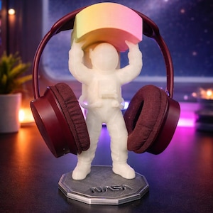 May include: A white astronaut figurine holding a rainbow-colored block, with maroon headphones draped over it. The figurine stands on a gray hexagonal base with "NASA" printed on it. The background is a blurred image of space.