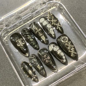 Könnte beinhalten: Ein Set schwarzer Kunstnägel mit silbernen und weißen Verzierungen. Die Nägel weisen verschiedene Designs auf, darunter Kreuze, Blumenmuster und Metallnieten. Die Nägel werden in einem transparenten Kunststoffbehälter präsentiert.