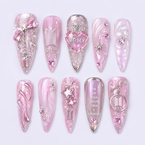 Op de afbeelding: Een verzameling van tien roze nail art ontwerpen. Elke nagel heeft een ander ontwerp, waaronder bedels, glitter en het BTS-logo. Sommige nagels hebben hart-, ster- of strikaccenten. De nagels zijn lang en puntig.