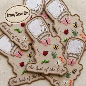 May include: A beige fabric patch featuring embroidered illustrations of a glass jar, a pink butterfly, a red ladybug, a green caterpillar, a yellow bee, and a green beetle. The text reads Iron/Sew On and the last of the bugs.