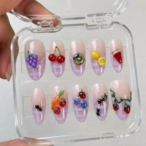 May include: A set of ten almond-shaped press-on nails in a clear plastic case. Each nail features a light purple grid pattern on a nude base, decorated with 3D charms including grapes, cherries, kiwi, lemon, watermelon, ants, carrots, tomatoes, blueberries, a ladybug, and strawberries.
