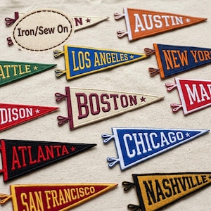 May include: A collection of embroidered felt pennant patches featuring various city names including Austin, Los Angeles, New York, Boston, Chicago, Atlanta, San Francisco, and Nashville. Each triangular patch includes the text Iron/Sew On.