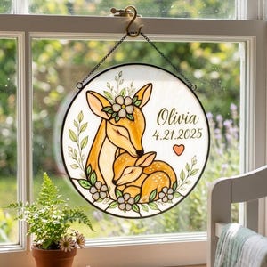 Puede incluir: Un adorno de ventana redondo de vidriera con un diseño de ciervo y cervatillo. La obra presenta una cierva abrazando a su cervatillo, rodeada de detalles florales. También incluye el nombre "Olivia" y la fecha "4.21.2025".