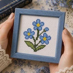 May include: A square, blue wooden frame containing a cross-stitch embroidery on white fabric. The embroidery depicts a cluster of blue forget-me-not flowers with yellow centers and green stems and leaves, centered within the frame.