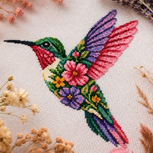 Op de afbeelding: Een kruissteekborduurwerk op witte stof met een kolibrie die is samengesteld uit levendige bloempatronen in roze, paarse, groene en gele tinten. De vogel heeft een lange zwarte snavel en vleugels gevuld met ingewikkelde, kleurrijke bloemmotieven.