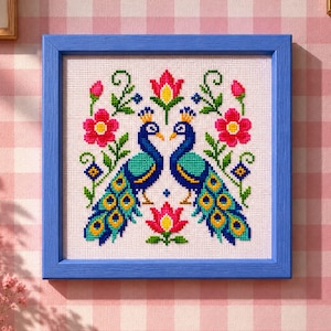 Puede incluir: Un bordado en punto de cruz cuadrado enmarcado en un borde de madera azul. El diseño presenta dos pavos reales simétricos de color azul y verde frente a frente, rodeados de flores rosas y rojas estilizadas, hojas verdes y pequeños detalles geométricos.