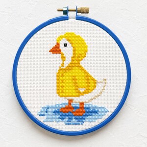May include: A cross-stitch embroidery piece featuring a yellow duckling wearing a yellow raincoat, standing in a blue puddle. The design is displayed within a circular blue plastic embroidery hoop with a metal tightening screw at the top.