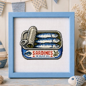 Pode incluir: Um bordado em ponto cruz quadrado numa moldura de madeira azul-clara com uma lata de sardinhas. O design mostra três peixes azuis dentro de uma lata aberta com o texto SARDINES IN OLIVE OIL e pequenas estrelas vermelhas e ícones de azeitona.