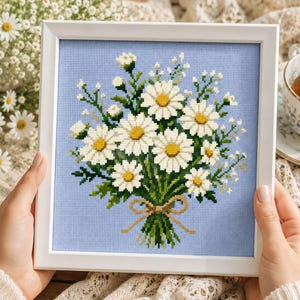 May include: A framed cross-stitch of a daisy bouquet. The daisies have white petals and yellow centers, tied with a brown ribbon against a light blue background. The frame is white.