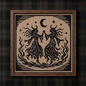 May include: A framed cross-stitch embroidery featuring two horned figures in long robes facing each other with hands touching. The design is rendered in black thread on a tan fabric background, surrounded by flames, stars, and a crescent moon.