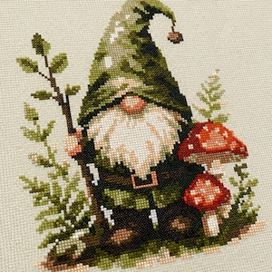 May include: A cross-stitch embroidery design on light beige fabric featuring a gnome wearing a tall, dark green pointed hat and a long white beard. The gnome holds a brown wooden staff and stands beside two red mushrooms with white spots.