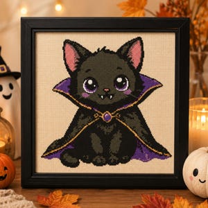 May include: A framed cross-stitch embroidery featuring a black cat with large purple eyes and small white fangs, wearing a black cape with a purple lining and a gold clasp with a purple gem, set against a light beige fabric background.