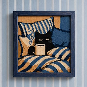 May include: A square cross-stitch embroidery in a dark blue wooden frame. The design features a black cat sitting among blue and beige striped pillows and blankets, holding a white mug. The cat has visible white eyes and whiskers.