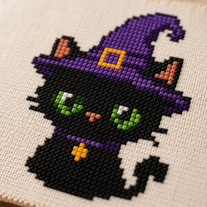 May include: A cross-stitch embroidery on beige fabric featuring a black cat wearing a purple witch hat with a yellow buckle. The cat has bright green eyes, a small pink nose, and a purple collar with a yellow star-shaped charm attached to it.