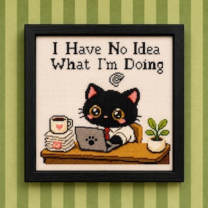 Puede incluir: Un bordado en punto de cruz enmarcado que muestra un gato negro sentado en un escritorio con un portátil, una pila de papeles, una taza de café y una pequeña planta en maceta. El texto sobre el gato dice I Have No Idea What I'm Doing.