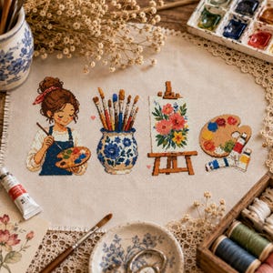 以下が含まれることがあります： ベージュの布地に施されたクロスステッチ刺繍。パレットを持って絵を描く人物、様々な絵筆が入った青と白の花柄の陶器製ジャー、花の絵が飾られたイーゼル、そして絵具のチューブが添えられた木製のアーティストパレットが描かれています。