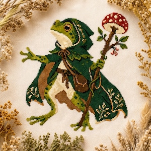 May include: A cross-stitch embroidery on cream fabric featuring a green frog wearing a dark green hooded cloak and a brown satchel. The frog holds a tall wooden staff topped with a red and white spotted mushroom, surrounded by dried foliage.