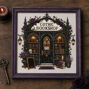 Op de afbeelding: Een ingelijst kruissteek borduurwerk met een winkelpui met de tekst GOTHIC BOOKSHOP en een bordje met OPEN. Het ontwerp bevat donkere architecturale details, paarse rozen, boekenplanken, een schedel, lantaarns en stapels boeken.