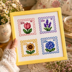 May include: A square cross-stitch embroidery in a yellow frame featuring four postage stamp designs. Each stamp depicts a different flower: a pink tulip labeled 10¢, purple lavender labeled 15¢, a yellow sunflower labeled 20¢, and a blue hydrangea labeled 25¢.