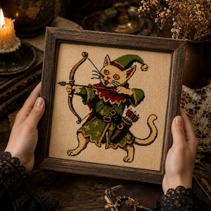 May include: A framed cross-stitch embroidery featuring a light-colored cat dressed in a green and red tunic with a matching pointed hat. The cat is depicted as an archer, holding a wooden bow and arrow, with a quiver of arrows strapped to its side.