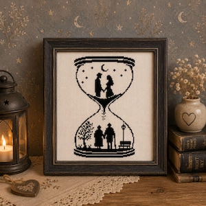 Könnte beinhalten: Eine gerahmte Kreuzstichstickerei mit einem schwarzen Silhouettenmotiv in Form einer Sanduhr. Der obere Teil zeigt ein junges Paar, das unter einer Mondsichel Händchen hält, während der untere Teil ein älteres Paar bei einem Baum zeigt.