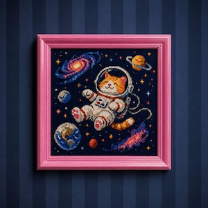 May include: A cross-stitch embroidery in a bright pink square frame featuring an orange and white cat wearing a white astronaut suit floating in deep space. The scene includes a spiral galaxy, a ringed planet, Earth, and small yellow stars.