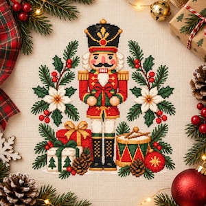 May include: A cross-stitch embroidery design featuring a central nutcracker figure in a red and gold uniform, flanked by green pine branches, white poinsettias, red berries, wrapped gift boxes, a drum, and pinecones on a light fabric background.