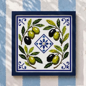 May include: A square cross-stitch embroidery framed in dark blue, featuring four olive branches with green and black olives arranged around a central blue and white geometric floral motif on a white background with blue corner accents.