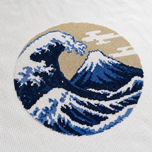May include: A circular cross-stitch embroidery on white fabric featuring a stylized ocean wave in shades of dark blue and light blue, with a mountain peak in the background against a tan circular sky area with white cloud shapes.