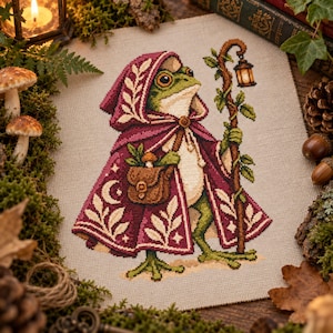 May include: A cross-stitch embroidery on beige fabric depicting a green frog wearing a deep magenta hooded cloak with white leaf patterns. The frog carries a brown satchel containing a mushroom and holds a wooden staff topped with a lantern.