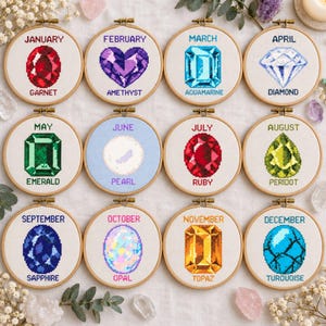 May include: Twelve circular wooden embroidery hoops featuring cross-stitch designs of birthstones for each month. Each hoop displays the month name and gemstone name in text, including Garnet, Amethyst, Aquamarine, Diamond, Emerald, Pearl, Ruby, Peridot, Sapphire, Opal, Topaz, and Turquoise.