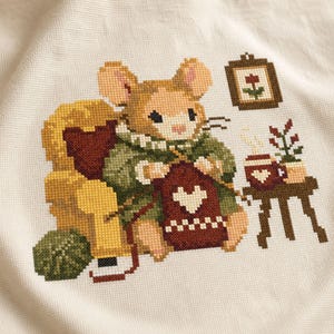 May include: A cross-stitch embroidery on cream fabric depicting a tan mouse sitting in a yellow armchair, knitting a dark red piece with a white heart. Beside the mouse is a small table with a steaming mug and a plant, plus a framed flower picture.