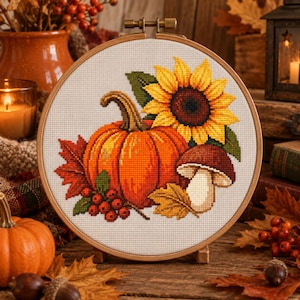 Könnte beinhalten: Ein runder Holzstickrahmen mit einem Kreuzstichmuster aus einem leuchtend orangefarbenen Kürbis, einer großen gelben Sonnenblume, einem braunen Pilz, roten Beeren und Herbstblättern auf einem weißen Stoff mit Gitterstruktur.
