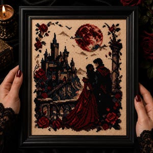 Op de afbeelding: Een ingelijst kruissteekkunstwerk dat een gotische scène uitbeeldt met een kasteel, een stel en een rode maan. Het kunstwerk heeft een zwart frame, rode rozen en een donker kleurenpalet. Het stel is gekleed in rode en zwarte kleding.