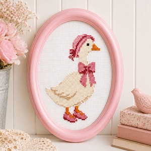 May include: A framed cross-stitch of a goose wearing a pink bonnet, bow, and shoes. The goose is cream-colored with an orange beak and black eyes. The pink frame is oval-shaped. The background is white.