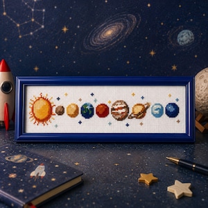 May include: A rectangular cross-stitch embroidery featuring the sun and eight planets in a horizontal row, framed in a dark blue border. The design is stitched on white fabric with small colourful crosses representing celestial bodies.