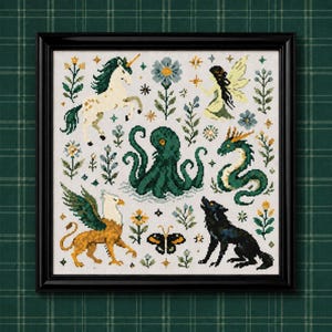 Puede incluir: Un bordado en punto de cruz enmarcado que presenta un unicornio blanco, un pulpo verde, un dragón verde, un grifo, un lobo negro, un hada y una mariposa, rodeados de varios motivos florales y pequeñas estrellas amarillas sobre un fondo de tela blanca.