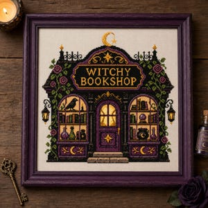 May include: A framed cross-stitch embroidery depicting a storefront with the text WITCHY BOOKSHOP. The design features a purple and black building with arched windows, shelves of books, a raven, a crystal ball, and a cauldron, all framed in purple.