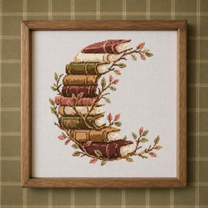 May include: A framed cross-stitch embroidery featuring a stack of books arranged in a crescent moon shape, intertwined with thin branches and small pink leaves. The books are rendered in shades of brown, green, and cream, set against a white fabric.