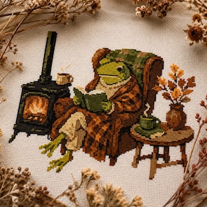 Könnte beinhalten: Eine Kreuzstichstickerei auf hellem Stoff, die einen grünen Frosch in einem braunen Sessel zeigt, der ein grünes Buch liest. Neben dem Sessel steht ein schwarzer Kaminofen mit Feuer, ein kleiner runder Tisch mit einer grünen Tasse und eine Vase.